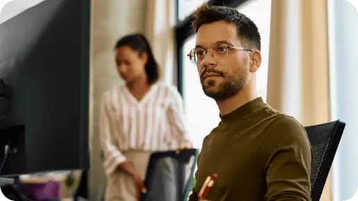 Employee seated at a desk facing a monitor while another person stands in the background.