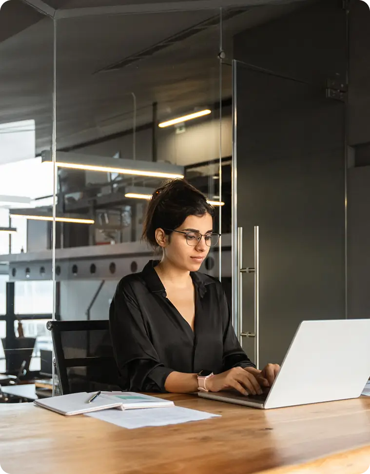 Woman wearing glasses working on a laptop in a glass-walled office.