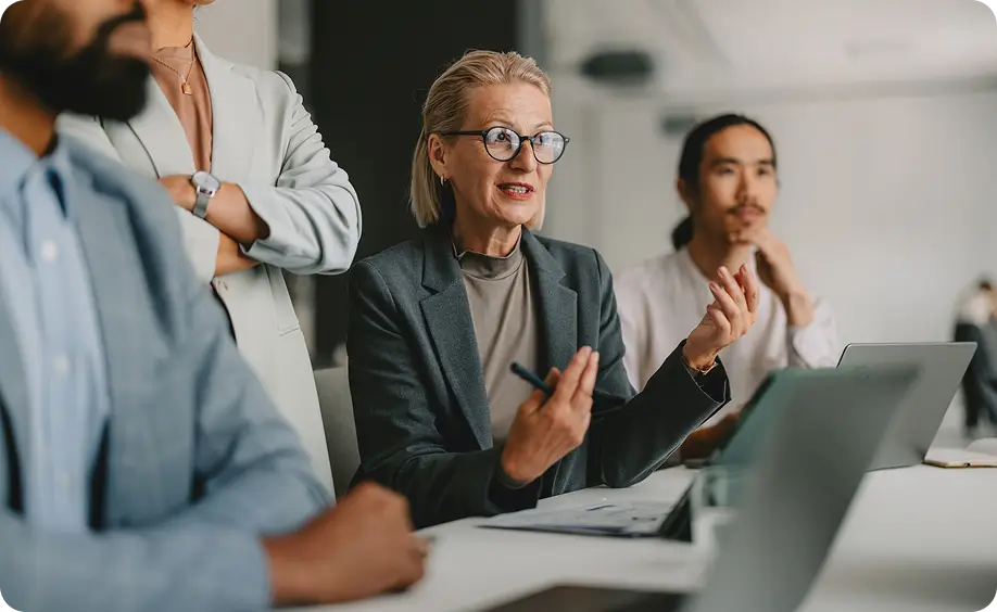 Group of coworkers seated at a table, with a woman speaking and gesturing during a meeting.