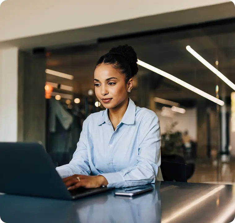 Woman in blue shirt seated at a table using a laptop with a smartphone placed beside it.