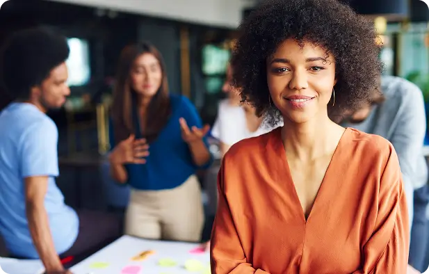 Woman smiling in an office setting with colleagues collaborating in the background.