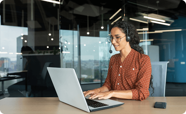 Woman seated at a desk working on a laptop with a headset in a glass-walled office.