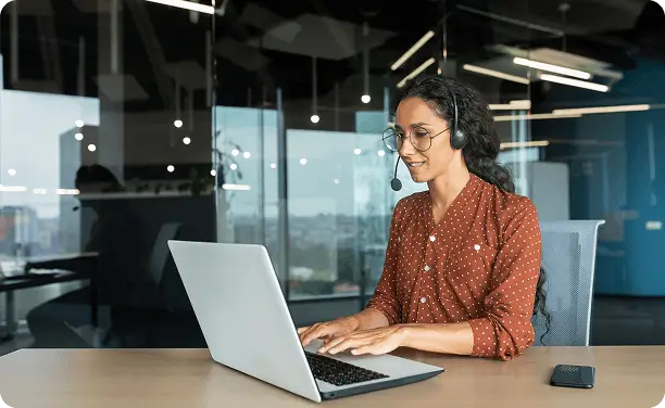 Woman seated at a desk working on a laptop with a headset in a glass-walled office.