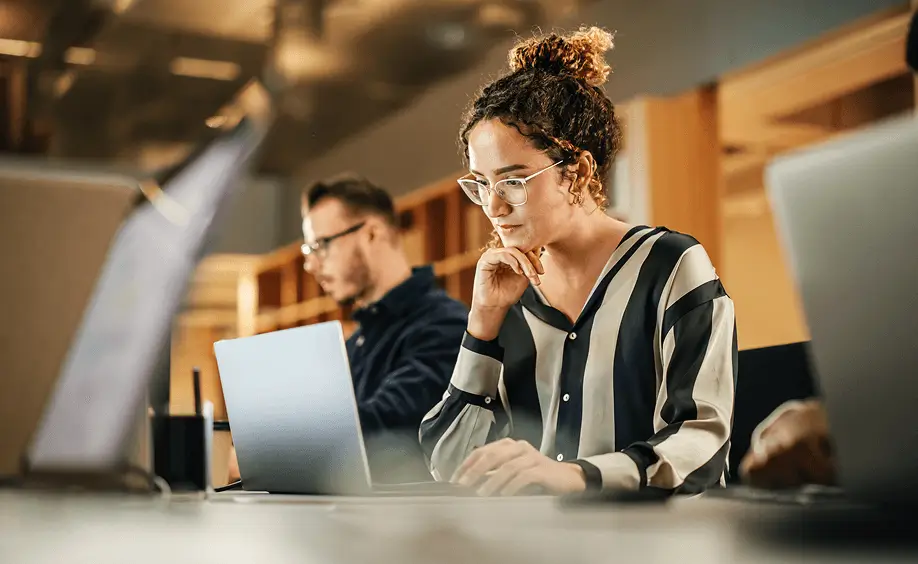 Two people using laptops at desks in a shared office workspace.