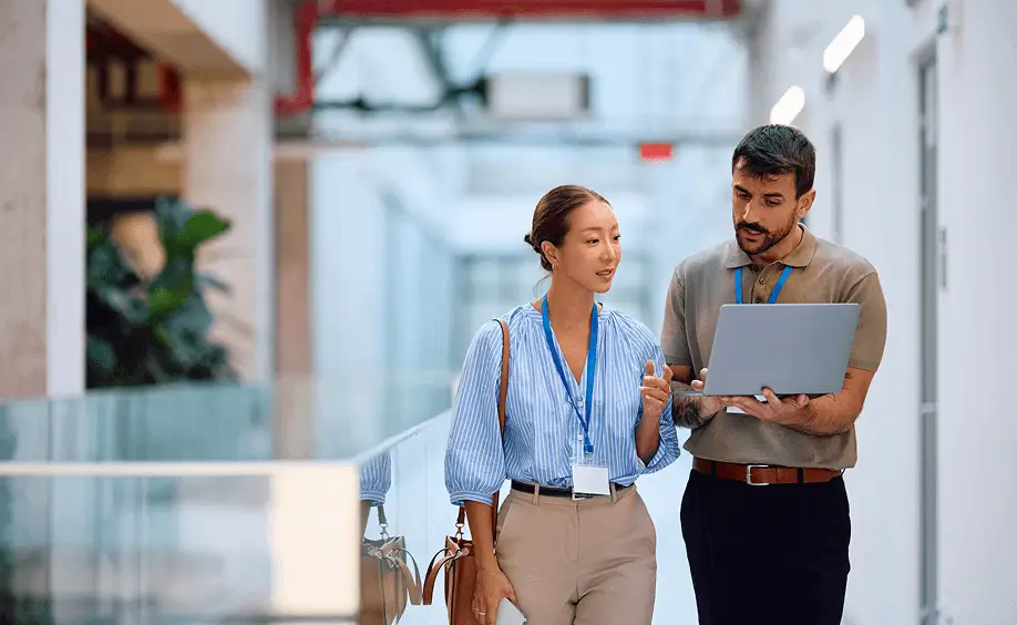 Two coworkers walking through an office hallway while reviewing information on a laptop.