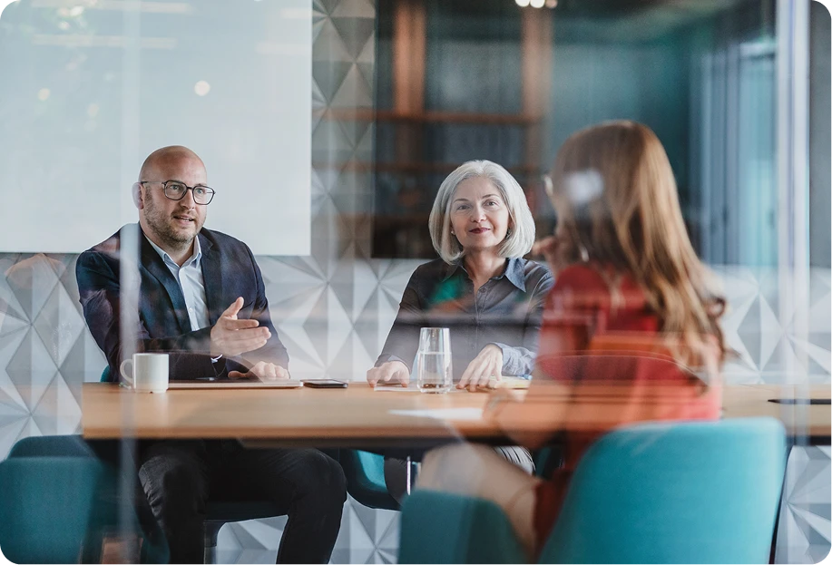 Three financial service professionals seated at a table in a glass-walled meeting room having a discussion.